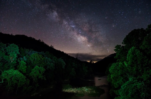 A starry night sky with the Milky Way over a dark forest and a calm river, illuminated by green light.