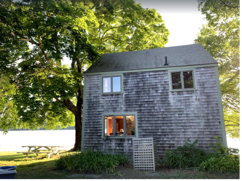 A two-story wooden house with a shingled exterior, surrounded by trees and near a lake, with a picnic table in the background.