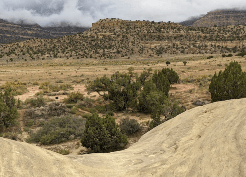 A vast desert landscape with rocky hills, sparse vegetation, and cloudy skies in the background.