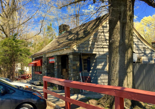 A quaint stone cottage with a sloped roof, surrounded by trees and a red railing, under a clear blue sky.
