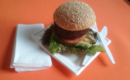 A sesame seed burger on a plate with lettuce, tomato, and pickles, accompanied by napkins and utensils on an orange background.