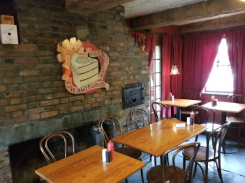 Cozy restaurant interior with wooden tables, brick wall, and a cake sign, featuring red curtains and condiments on tables.