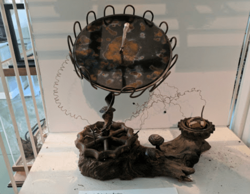 A unique sculpture featuring a circular metal clock atop a twisted base with organic shapes and textures.
