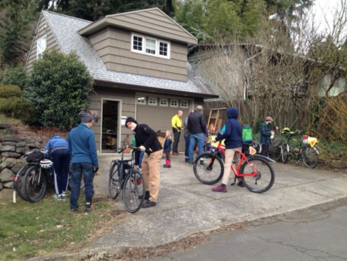 A group of people working on bicycles outside a house, with trees and a garage in the background.