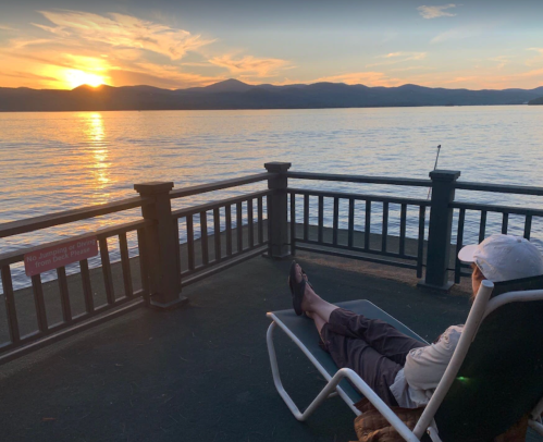 A person relaxes in a chair on a deck, watching a sunset over a calm lake and distant mountains.