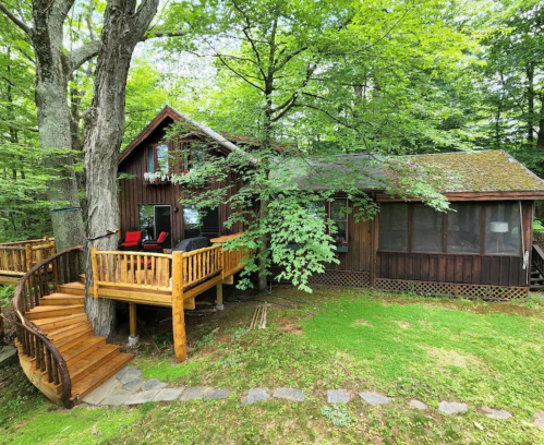 A rustic wooden cabin surrounded by lush green trees, featuring a deck with seating and a winding staircase.