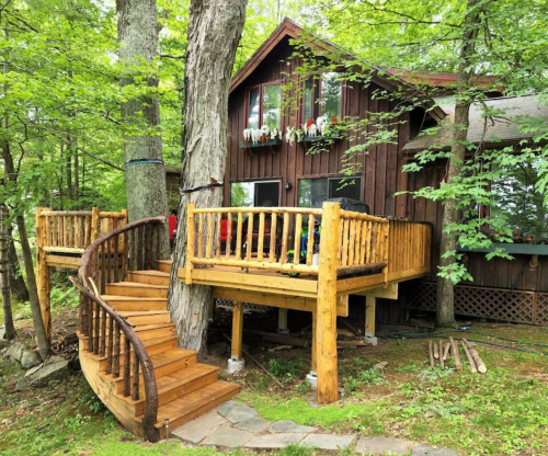 A rustic wooden cabin surrounded by trees, featuring a spiral staircase and a deck with railings.