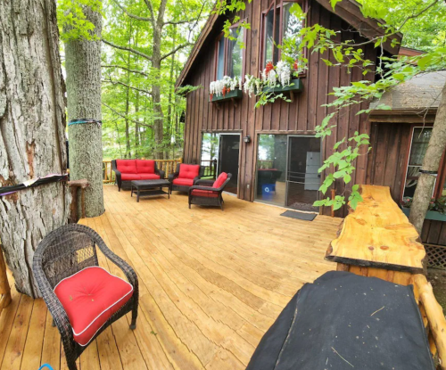 A wooden deck with red chairs and a table, surrounded by trees and a rustic cabin in the background.