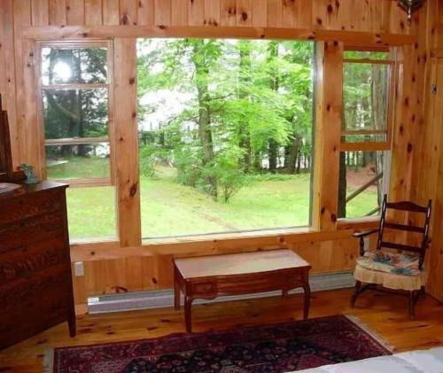 Cozy wooden room with a large window overlooking a green outdoor landscape and a small table in front.