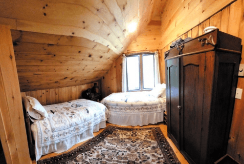 Cozy attic bedroom with two beds, wooden walls, and a vintage wardrobe, illuminated by natural light from a window.