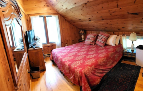 Cozy attic bedroom with wooden walls, a large bed, decorative pillows, a TV, and a lamp beside a window.