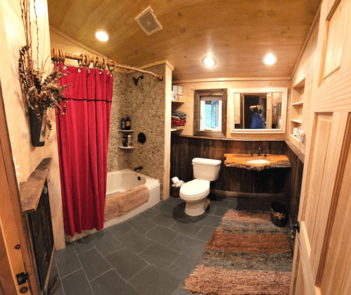 Cozy bathroom featuring a red shower curtain, stone shower, wooden accents, and a rustic rug.