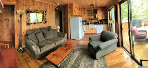 Cozy wooden cabin interior with a sofa, armchair, kitchen area, and large windows overlooking a deck.