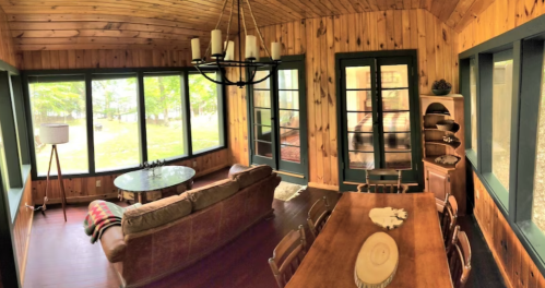 Cozy sunroom with wooden walls, a sofa, dining table, and large windows overlooking a green landscape.