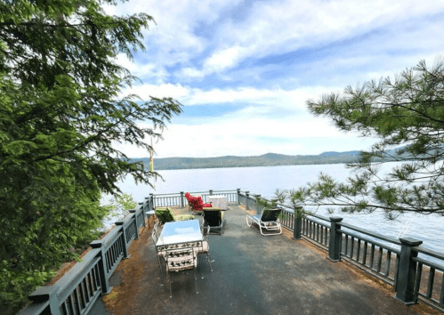 A scenic lakeside deck with chairs and tables, surrounded by trees and mountains under a blue sky.