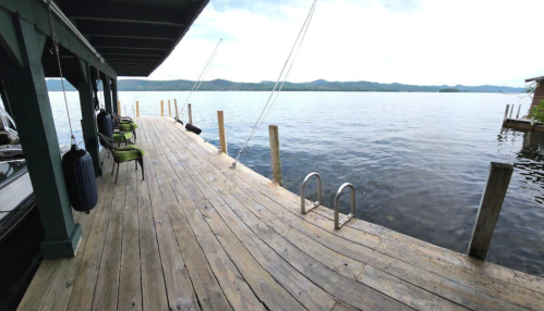 A wooden dock extends over calm water, with green chairs and mountains in the background under a cloudy sky.