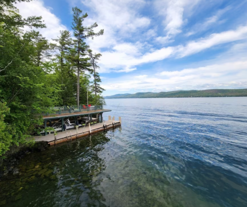 A serene lakeside view with a wooden dock, surrounded by trees and mountains under a partly cloudy sky.