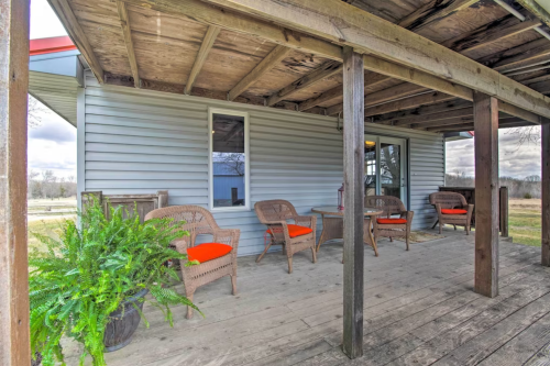 A porch with wooden flooring and wicker chairs featuring orange cushions, surrounded by greenery and a rural landscape.