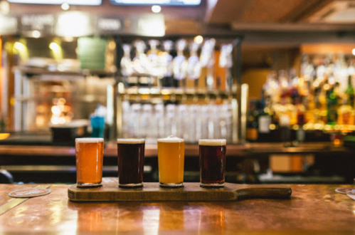 A wooden flight board with four different beers in glasses, set on a bar with a blurred background of bottles.