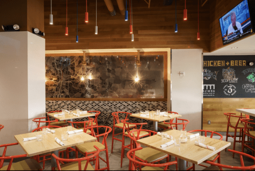 Modern restaurant interior with wooden walls, colorful pendant lights, and red chairs around wooden tables.