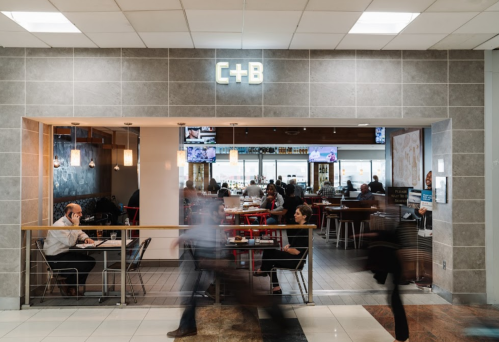 A busy airport restaurant with a modern design, featuring patrons dining and a bar area in the background.