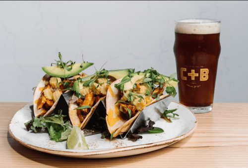 Three tacos filled with shrimp and avocado, served on a plate with a side salad and a glass of beer.