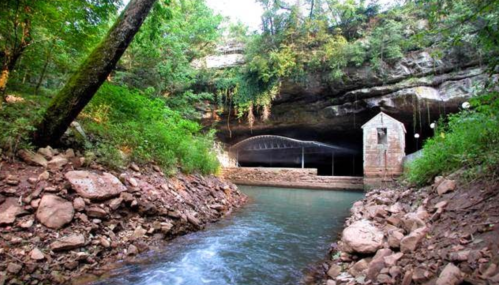 A serene river flows past a cave entrance, surrounded by lush greenery and rocky terrain.