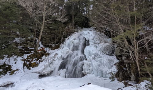 A frozen waterfall surrounded by snow-covered rocks and trees in a winter landscape.