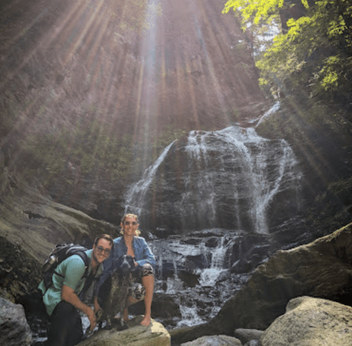 Two people and a dog pose by a waterfall, surrounded by lush greenery and sunlight streaming through the trees.