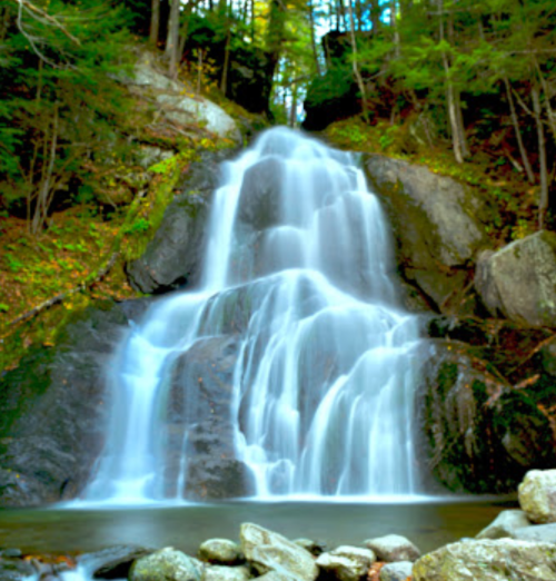 A serene waterfall cascading over rocks, surrounded by lush green trees and a tranquil pool at the base.