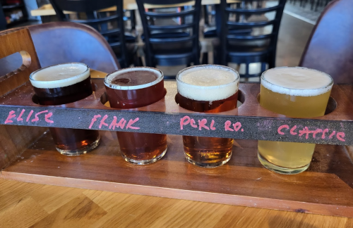 A wooden flight holder with four different beer samples, each labeled with names, on a table in a bar setting.
