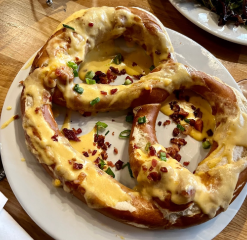 A large pretzel topped with melted cheese, bacon bits, and green onions, served on a white plate.