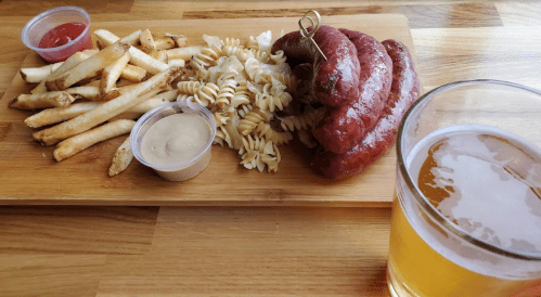 A wooden platter with fries, pasta, sausages, dipping sauces, and a glass of beer.