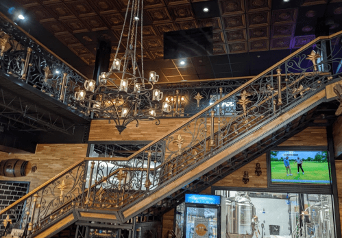 A stylish interior featuring a staircase with ornate ironwork, a chandelier, and a TV screen in a warm, wooden setting.