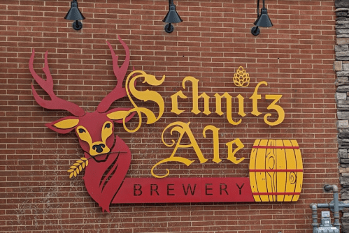 Colorful sign for "Schnitz Ale Brewery" featuring a deer with antlers and a barrel against a brick wall.