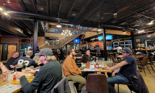 A lively restaurant interior with patrons at tables, a bar area, and a staircase in the background. Warm lighting and a casual vibe.