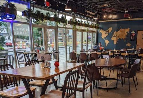 A cozy restaurant interior with wooden tables, chairs, and large windows, featuring plants and a world map on the wall.