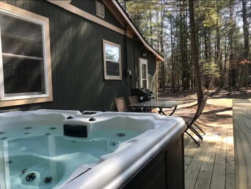 A hot tub on a wooden deck beside a cabin, surrounded by trees in a serene outdoor setting.