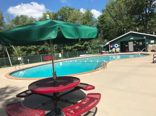 A swimming pool surrounded by trees, featuring a green umbrella over a red picnic table.