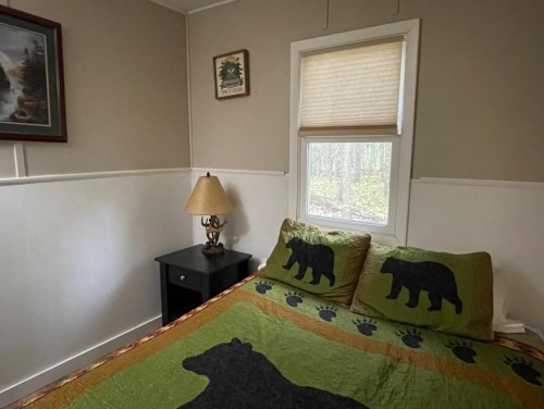Cozy bedroom with a bear-themed quilt, a bedside lamp, and a window overlooking greenery.