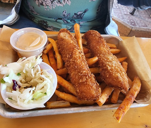 A tray of golden-brown fish fillets on a bed of fries, accompanied by coleslaw and a small cup of dipping sauce.