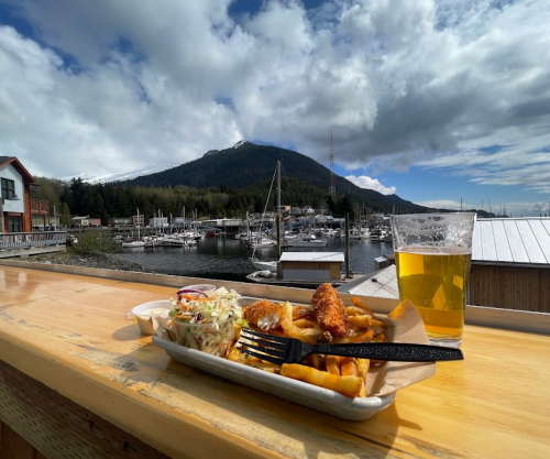 A plate of fish and chips with coleslaw and a beer, overlooking a marina and mountains under a cloudy sky.