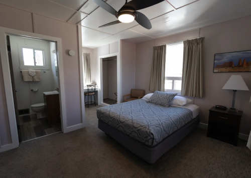 A cozy bedroom with a bed, nightstand, and a view of a bathroom and a small workspace in the background.