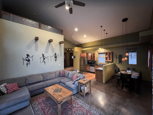 Cozy living area with a gray sofa, wooden table, and kitchen, featuring decorative wall art and warm lighting.