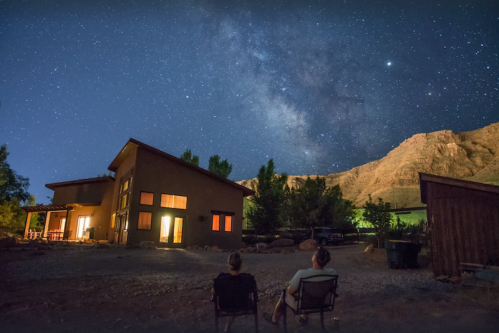 Two people sit outside a house, gazing at a starry night sky filled with the Milky Way and surrounding mountains.