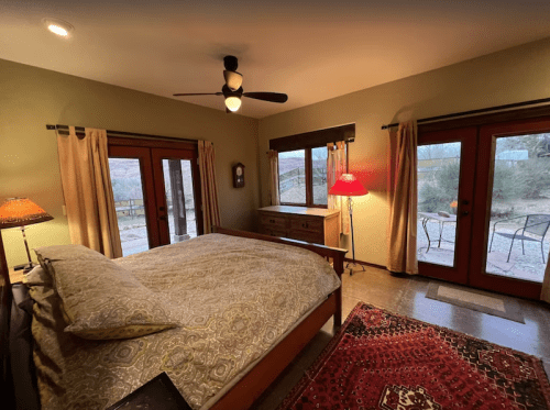 Cozy bedroom with a large bed, wooden furniture, and doors leading to an outdoor patio. Warm lighting and a rug add comfort.