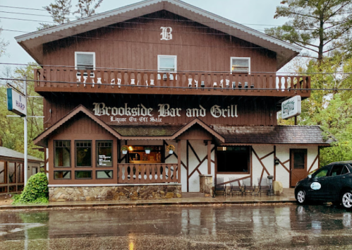 A brown wooden building with "Brookside Bar and Grill" sign, surrounded by trees, on a rainy day.