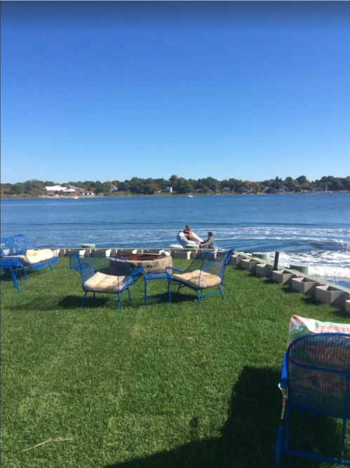 A sunny lakeside scene with blue chairs on grass, and two people in a small boat on the water.