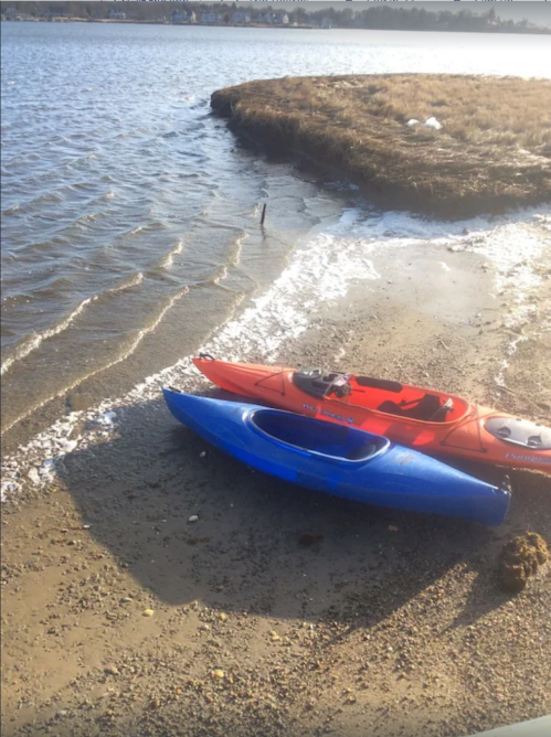 Two kayaks, one red and one blue, are resting on a sandy shore by a calm waterway with gentle waves.