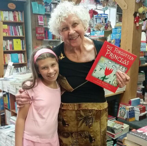 An elderly woman and a young girl smile together, holding a book titled "The Forgotten Princess" in a bookstore.